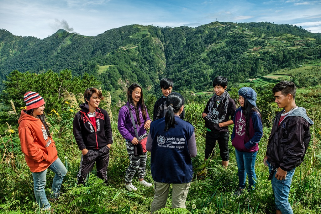 A WHO staff member stands, with their back to the camera, in the middle of a group of smiling teenagers in Benguet, Philippines. Benguet’s lush highlands can be seen in the background.